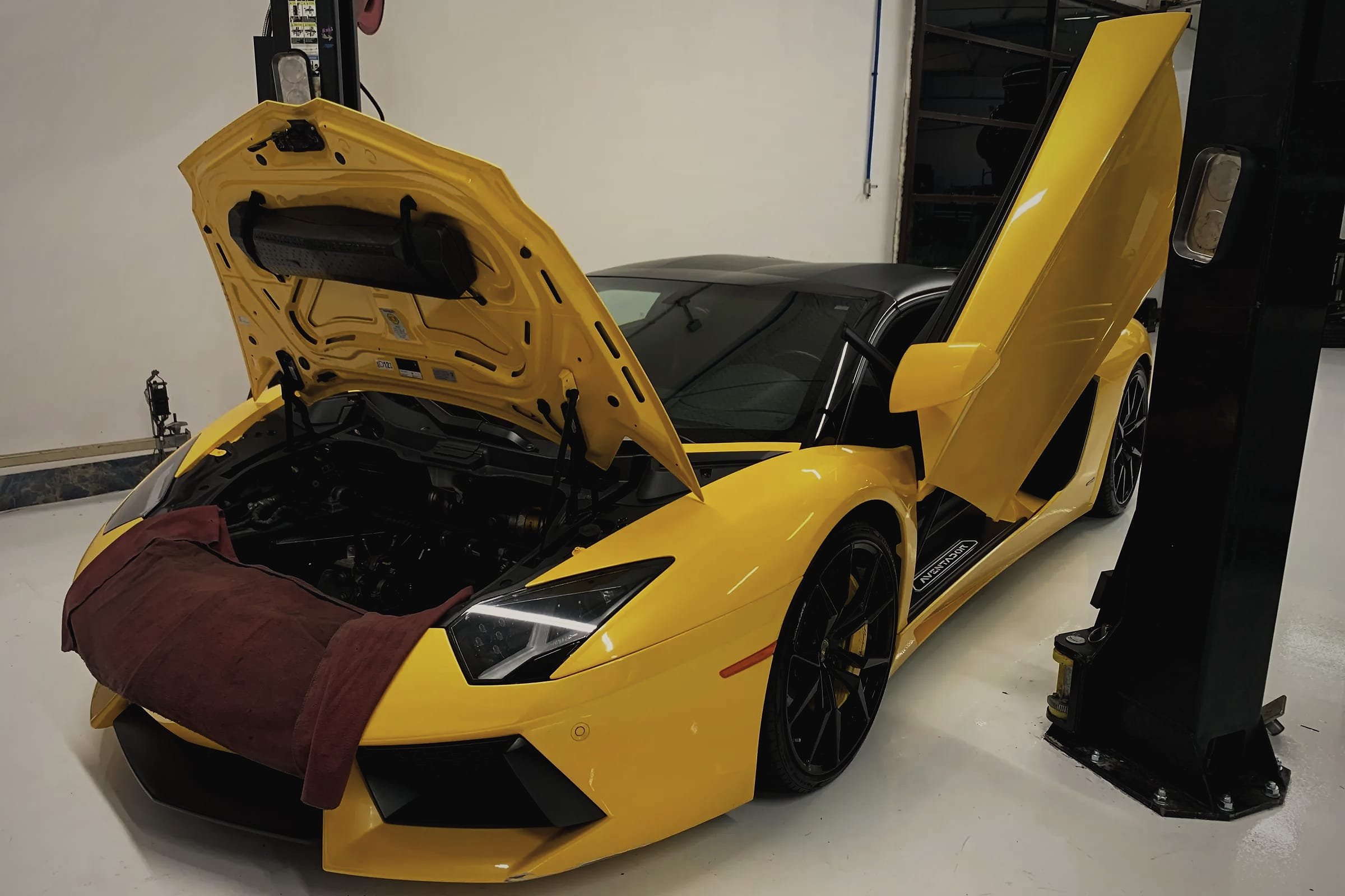 A yellow Lamborghini Aventador Roadster with its front bonnet and a scissor door open and a fender cover over the nose, beside a two-post lift.