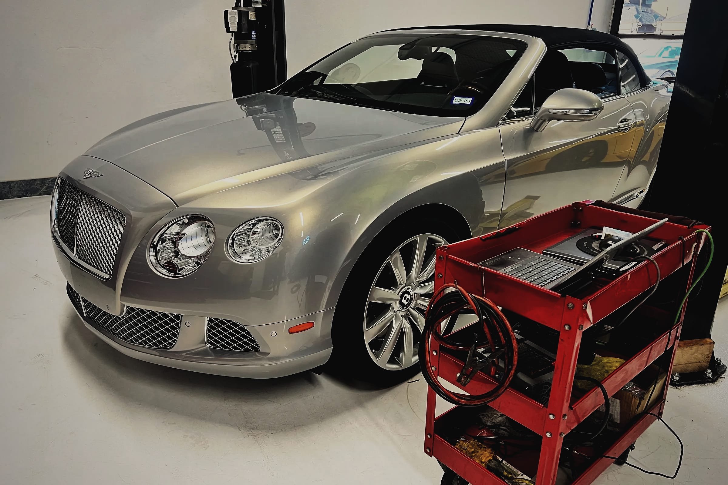 A silver Bentley Continental GTC convertible in the workshop, with a tool cart and charging leads beside it.