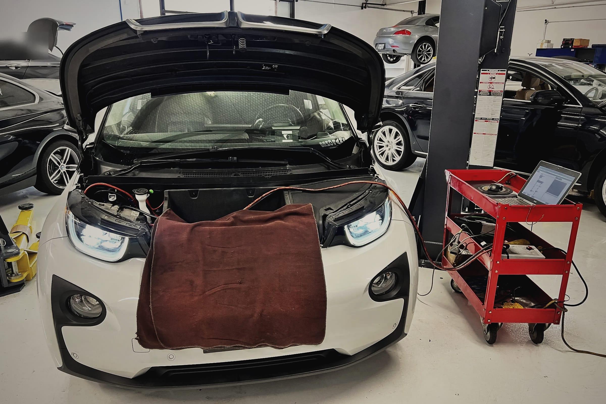A white BMW i3 with its front cover open and a fender cover over the nose, cabled to a tool cart with a laptop.