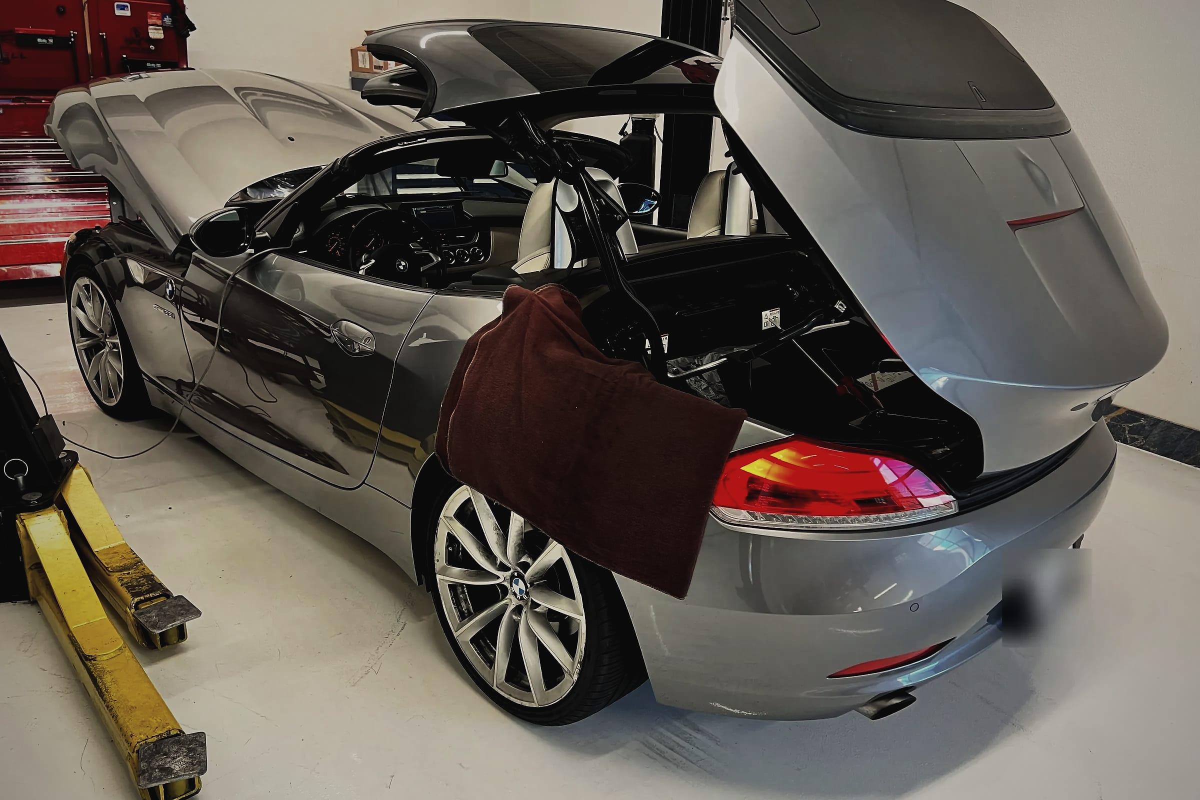 A grey BMW Z4 roadster with its folding hard top part way open and the boot lid raised, a fender cover over the rear wing.