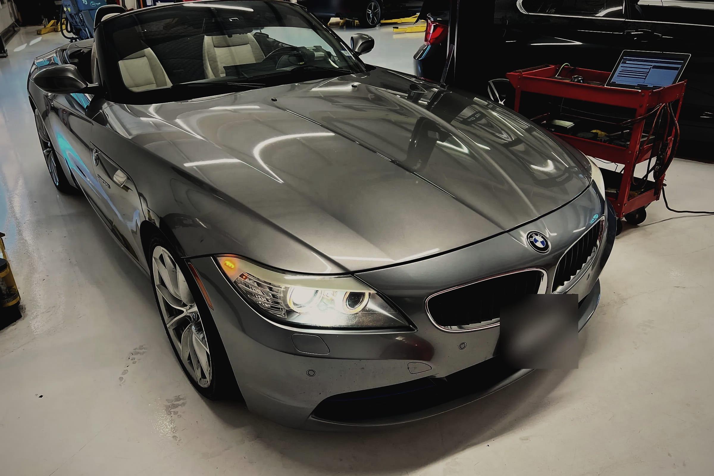 A grey BMW Z4 roadster with the roof down on the workshop floor, a tool cart with a laptop beside it.