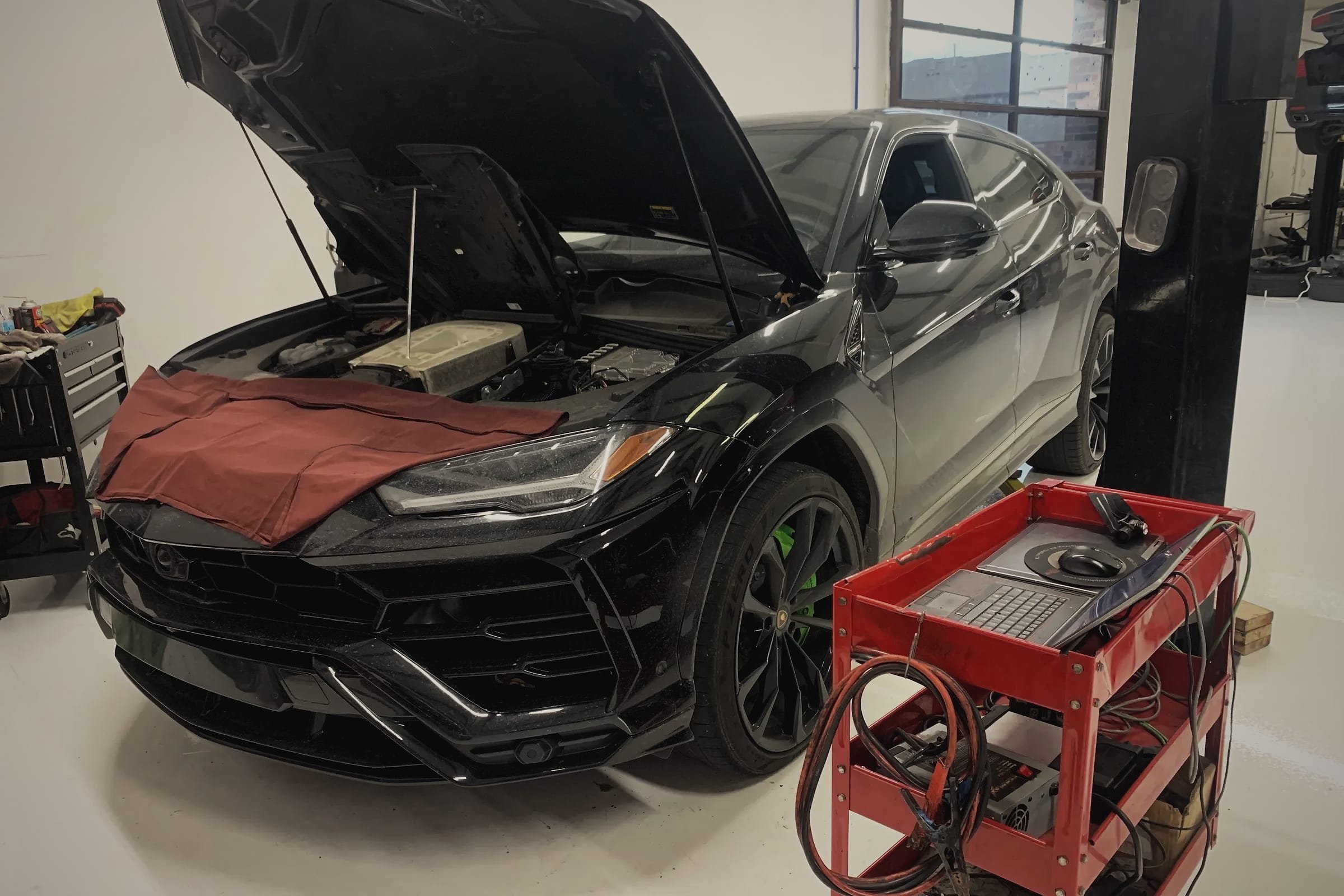 A black Lamborghini Urus with its bonnet open and a fender cover over the front, next to a tool cart with charging leads.