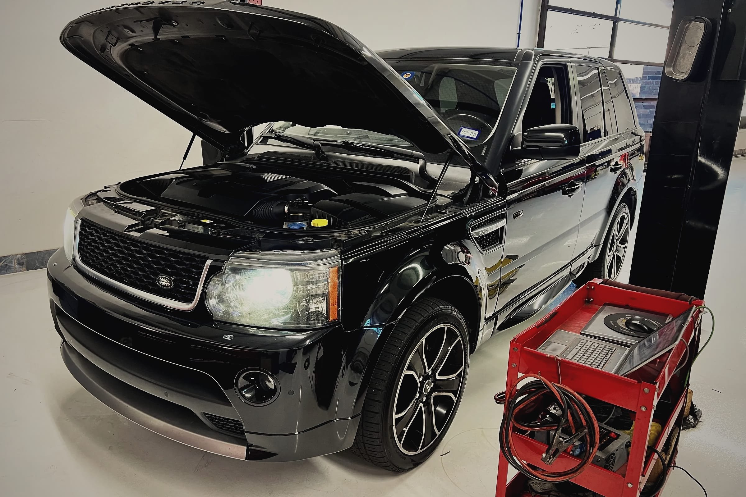 A black Range Rover Sport with its bonnet open beside a two-post lift, a tool cart with charging leads next to it.