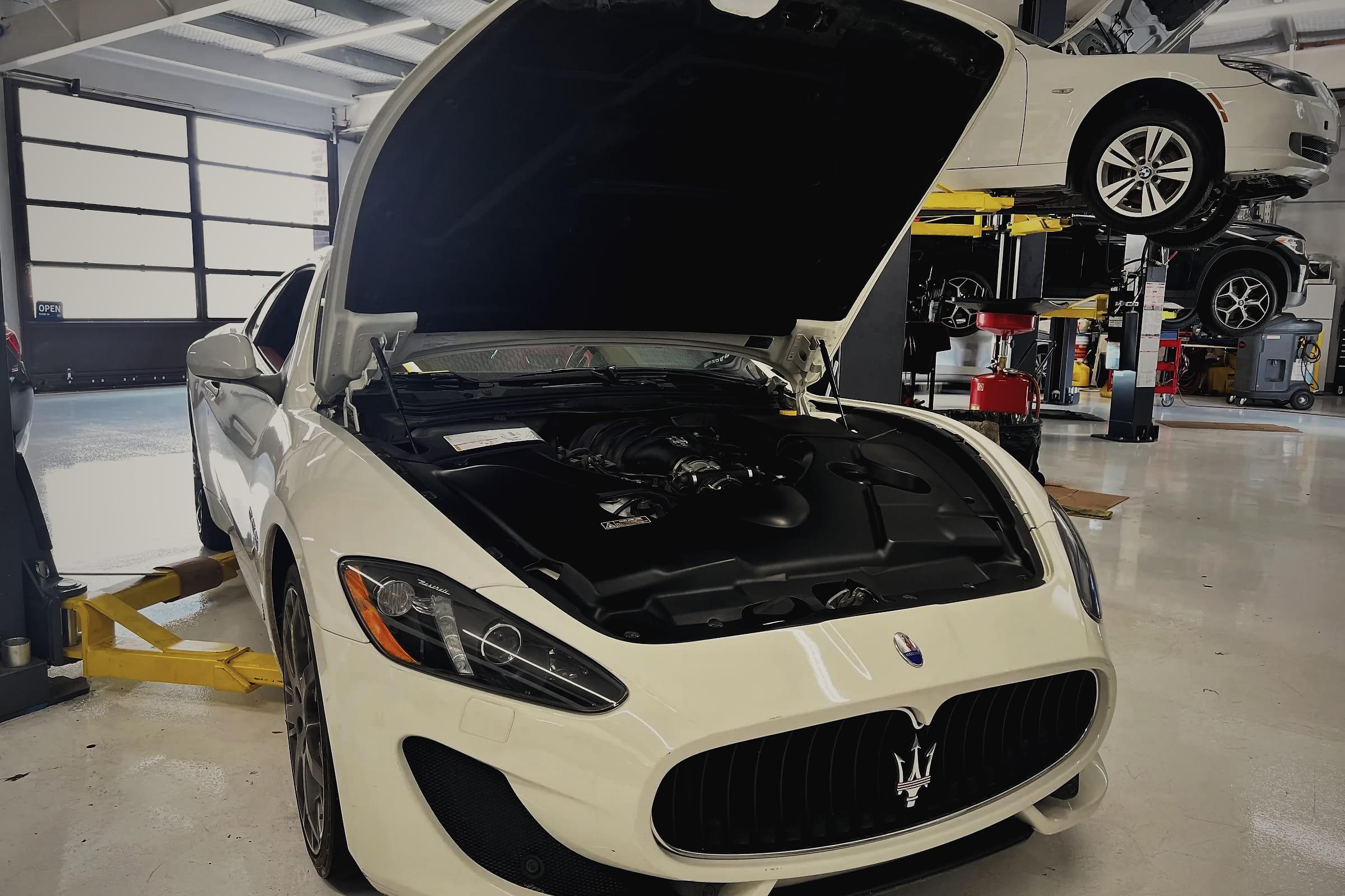 A white Maserati GranTurismo with its bonnet open showing the V8, a BMW raised on a lift behind it.