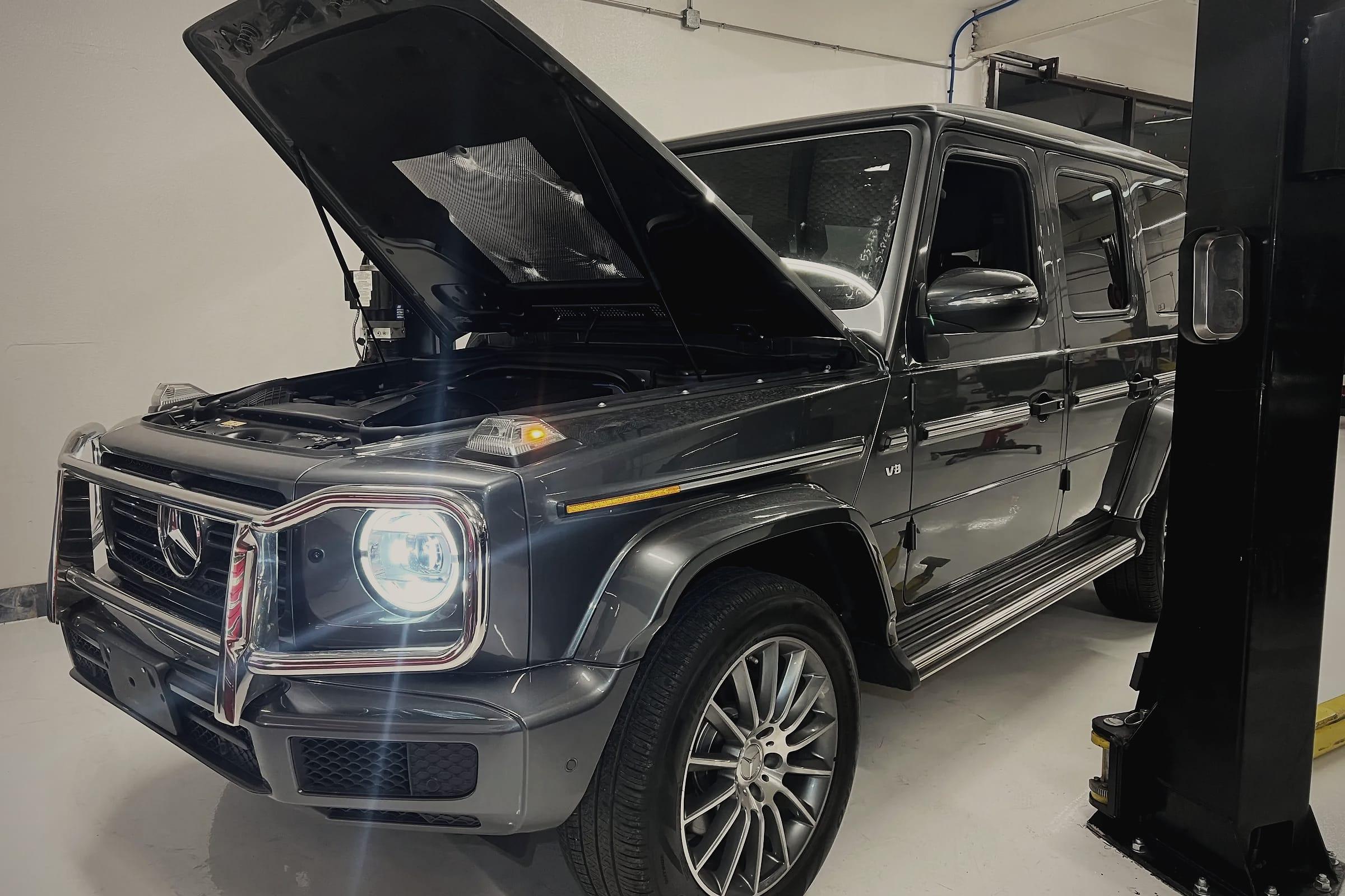 A grey Mercedes-Benz G-Class with its bonnet open beside a two-post lift in the workshop.