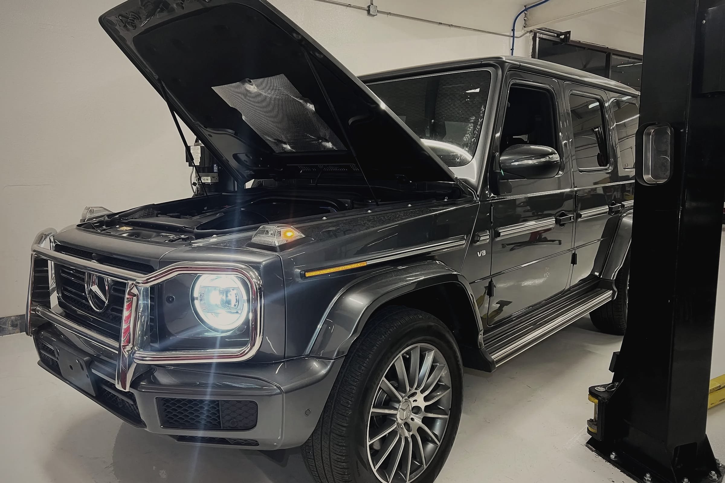 A grey Mercedes-Benz G-Class with its bonnet open beside a two-post lift in the workshop.