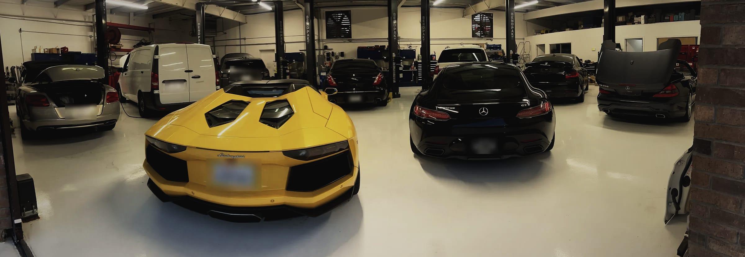 A wide view of the workshop: a yellow Lamborghini Aventador Roadster and a black Mercedes-AMG GT seen from behind, with a Bentley, a van and more cars between the lifts.